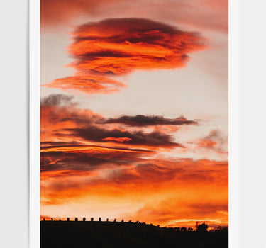 A vertical photographic art print of a dramatic burning sunset sky with glowing orange and red clouds, featuring a dark silhouette of a fence and small structures along the horizon. 