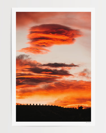 A vertical photographic art print of a dramatic burning sunset sky with glowing orange and red clouds, featuring a dark silhouette of a fence and small structures along the horizon. 